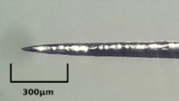 A metal needle tip comes to a point against a white background. A scale bar in the lower left shows a 300 micrometer length.